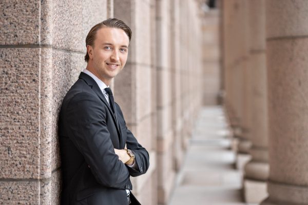 Ilmari Nurminen hymyilee ja seisoo nojaten seinään eduskuntatalon pylväiden lomassa kädet puuskassa. Hänellä on päällään musta puku valkoisella paidalla ja mustalla kravaatilla.
